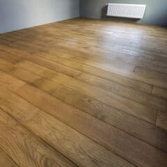 An empty room with wooden flooring, light gray walls, and a white radiator mounted on the wall. The room is lit by natural light, highlighting the smooth texture and warm tones of the wooden floorboards.