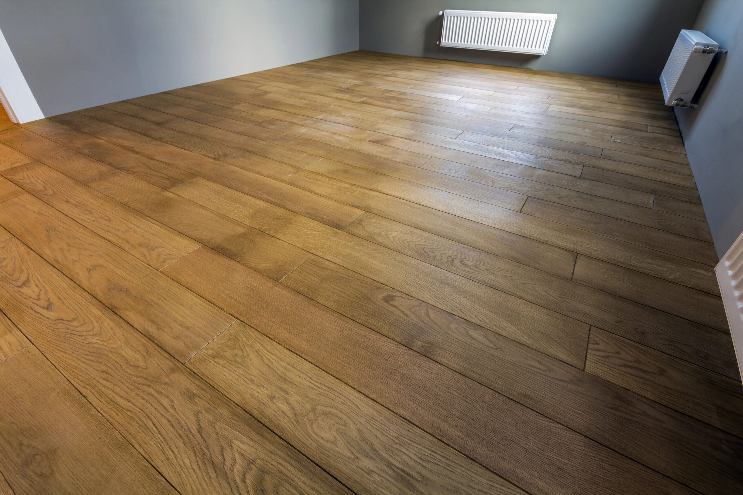 An empty room with wooden flooring, light gray walls, and a white radiator mounted on the wall. The room is lit by natural light, highlighting the smooth texture and warm tones of the wooden floorboards.