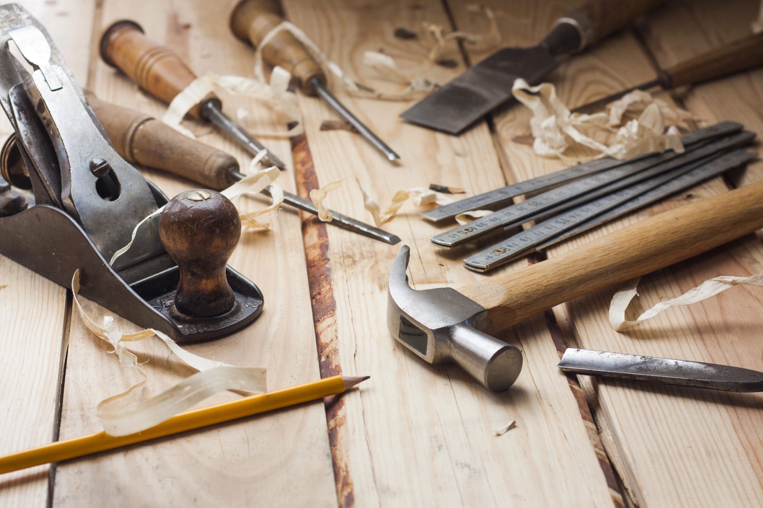 A variety of woodworking tools spread out on a wooden surface. Items include a hand plane, chisels, a hammer, files, and shavings of wood, along with a pencil. The scene suggests an ongoing woodworking project.