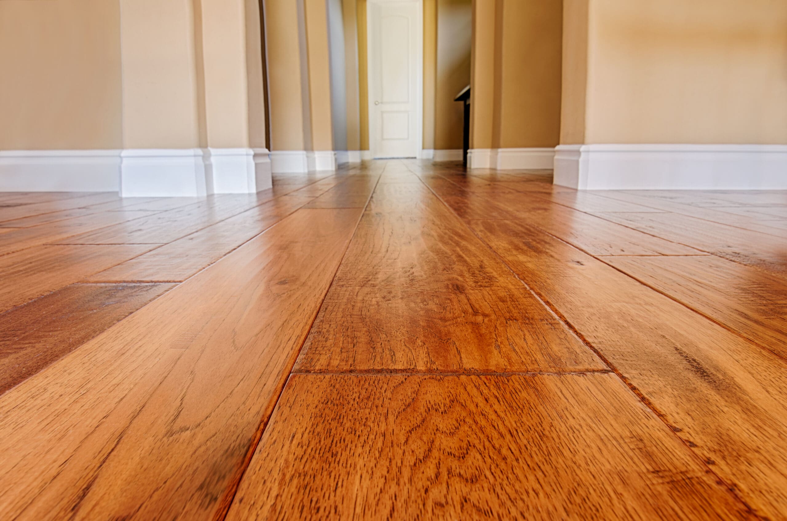 A close-up view of a polished wooden floor in a hallway with beige walls and white baseboards. In the background, an open door leads to another room, and light casts soft shadows across the surface, emphasizing the wood grain texture.