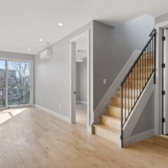 Modern open-concept living room with light wood flooring and neutral grey walls. A staircase with black railing leads to an upper level. Large sliding glass doors at the end provide natural light and a view of trees. A door under the stairs opens to a small room.