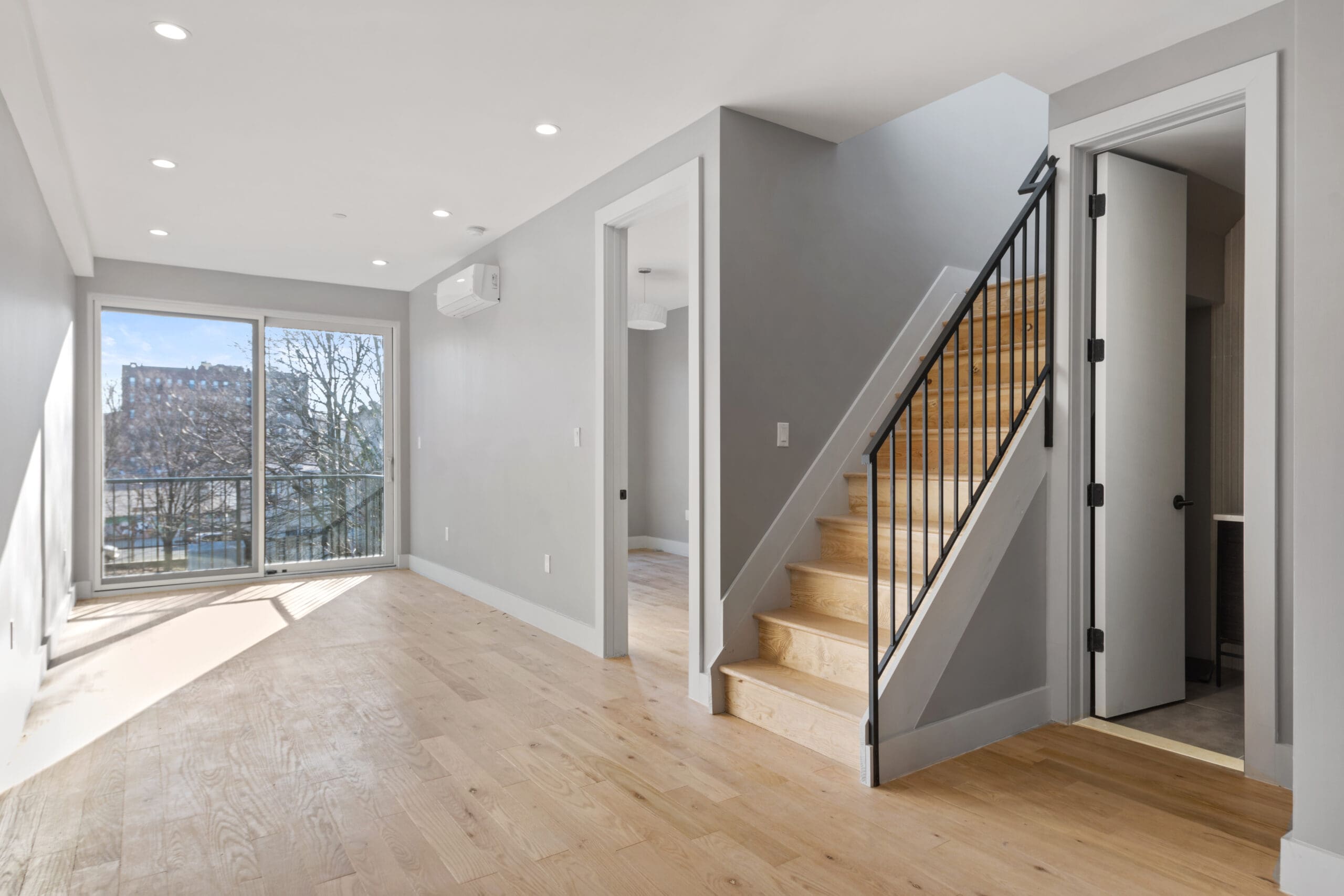 Modern open-concept living room with light wood flooring and neutral grey walls. A staircase with black railing leads to an upper level. Large sliding glass doors at the end provide natural light and a view of trees. A door under the stairs opens to a small room.