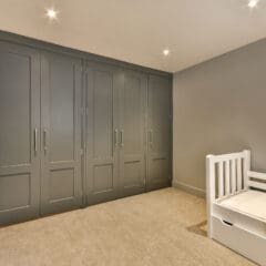 A minimalist bedroom featuring a white child’s bed with storage drawers underneath on the right, and a wall of large, gray built-in wardrobes with panel doors and modern handles on the left. The room has beige carpeting and is lit by recessed ceiling lights.