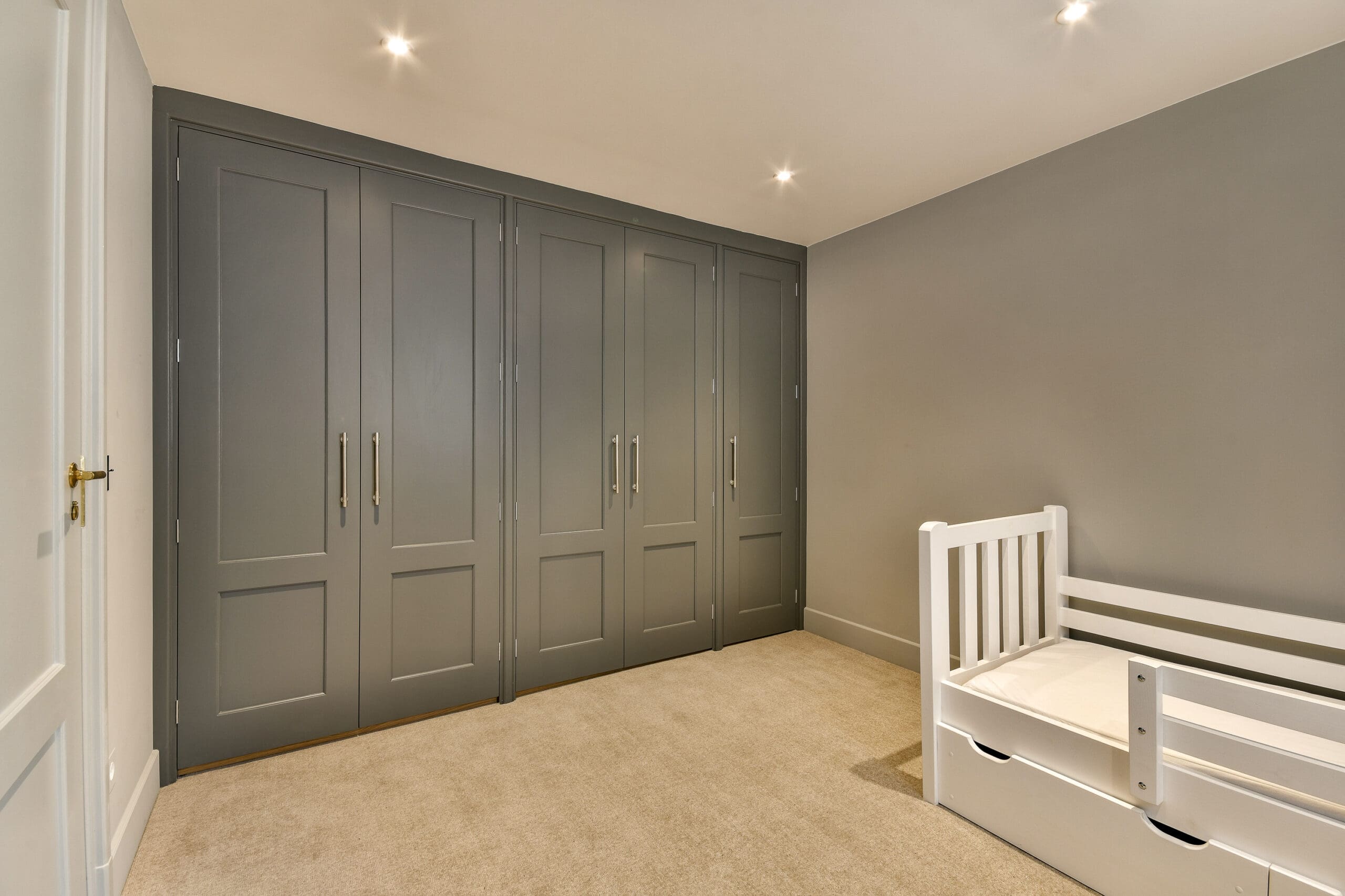 A minimalist bedroom featuring a white child’s bed with storage drawers underneath on the right, and a wall of large, gray built-in wardrobes with panel doors and modern handles on the left. The room has beige carpeting and is lit by recessed ceiling lights.