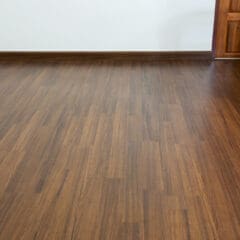 A room with polished wooden flooring, light-colored walls, and a wooden door on the right side. The floor has a smooth, shiny finish with a rich, natural wood grain pattern.