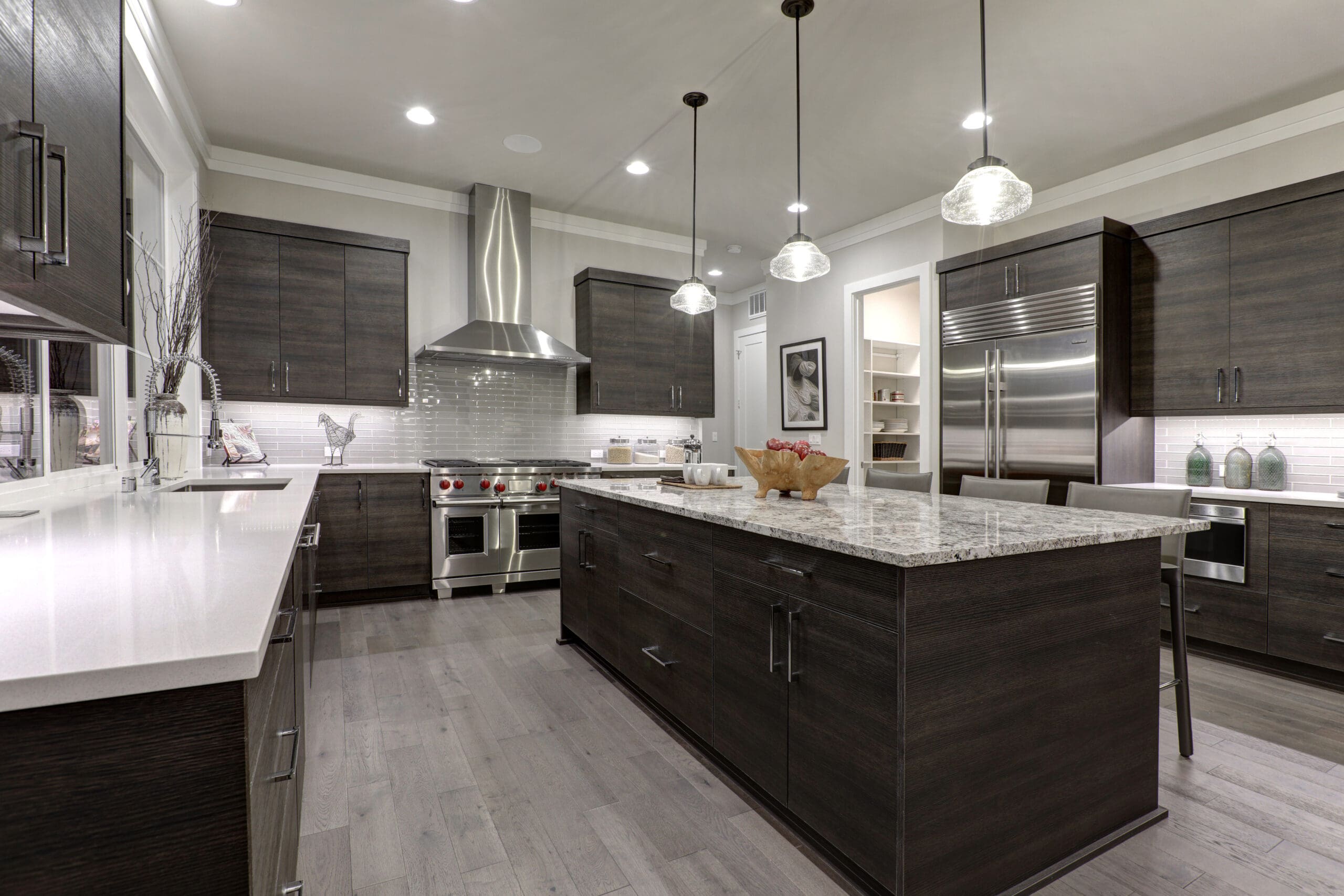 Modern kitchen with dark wood cabinets, stainless steel appliances, and a large island with a light granite countertop. Three pendant lights hang above the island. The kitchen features a gas cooktop with a hood and recessed lighting in the ceiling.