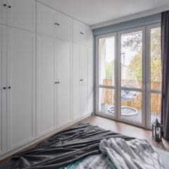 A bright bedroom with a large window and glass door opening to a balcony. The room features white built-in wardrobes along one wall, a bed with rumpled gray and white bedding, and dark curtains. On the balcony, there are hanging chairs and a lantern.