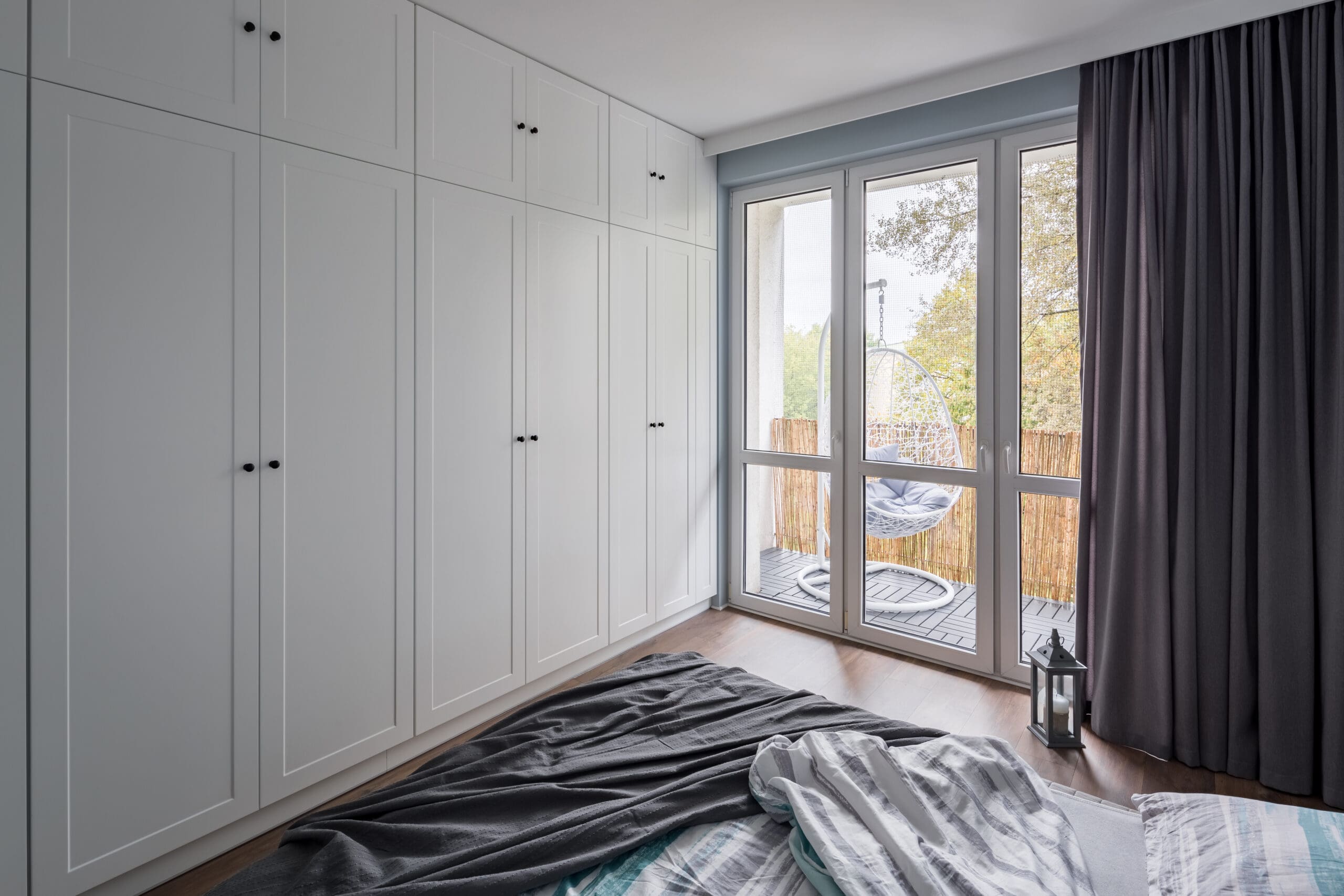 A bright bedroom with a large window and glass door opening to a balcony. The room features white built-in wardrobes along one wall, a bed with rumpled gray and white bedding, and dark curtains. On the balcony, there are hanging chairs and a lantern.