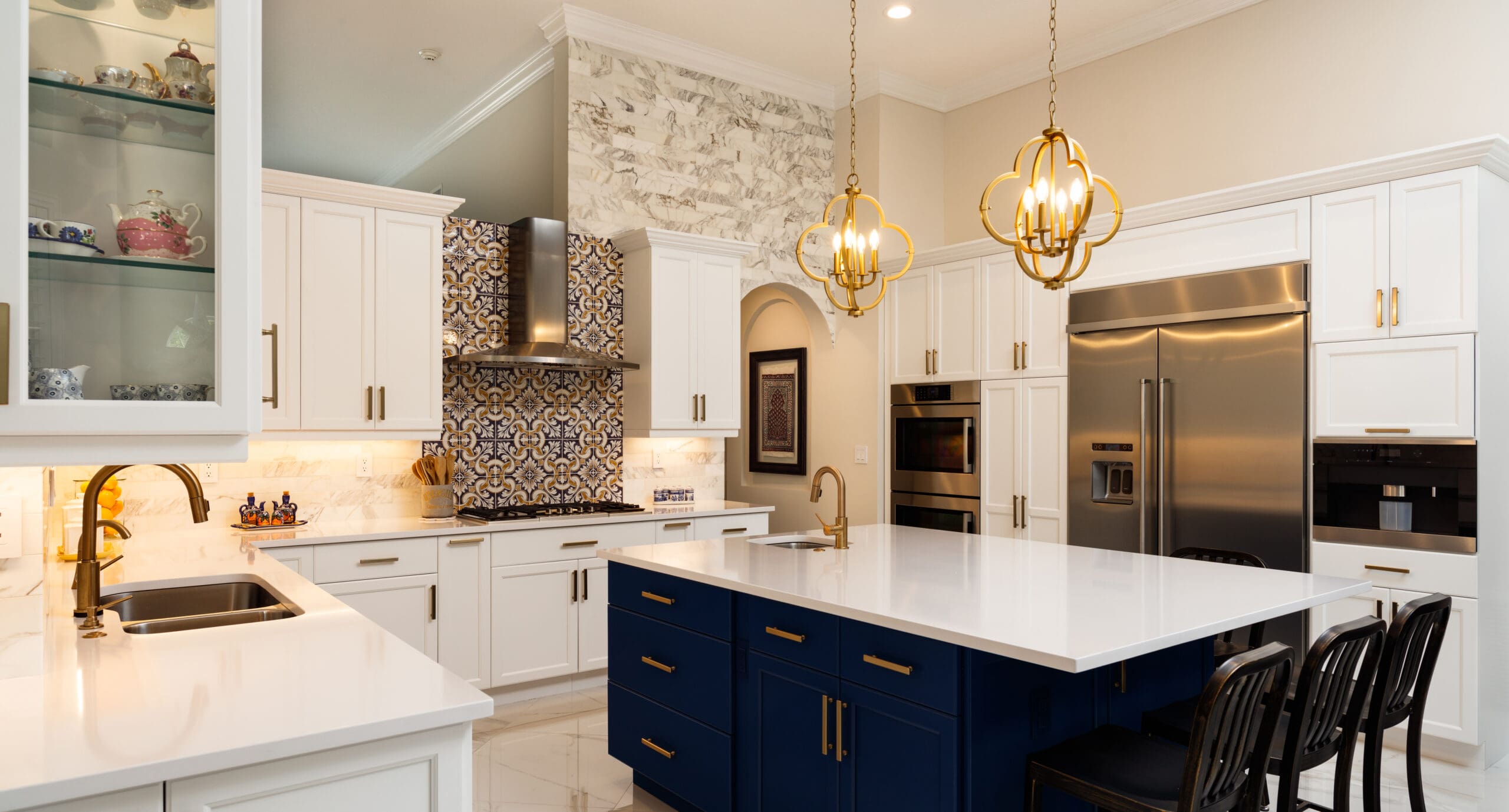 A modern kitchen with white cabinets, a large white island with a dark blue base, and brass fixtures. The back wall features a decorative tile backsplash behind a stainless steel stove. Two gold pendant lights hang over the island, and a stainless steel refrigerator is on the right.