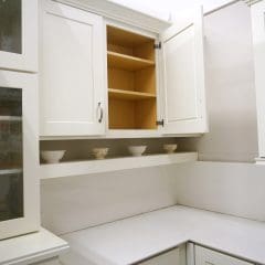 A white kitchen features upper cabinets with stainless steel handles, an open cabinet showing empty wooden shelves, a beige marble countertop, and decorative bowls on a ledge beneath the open cabinet. The walls have a matching beige marble backsplash.