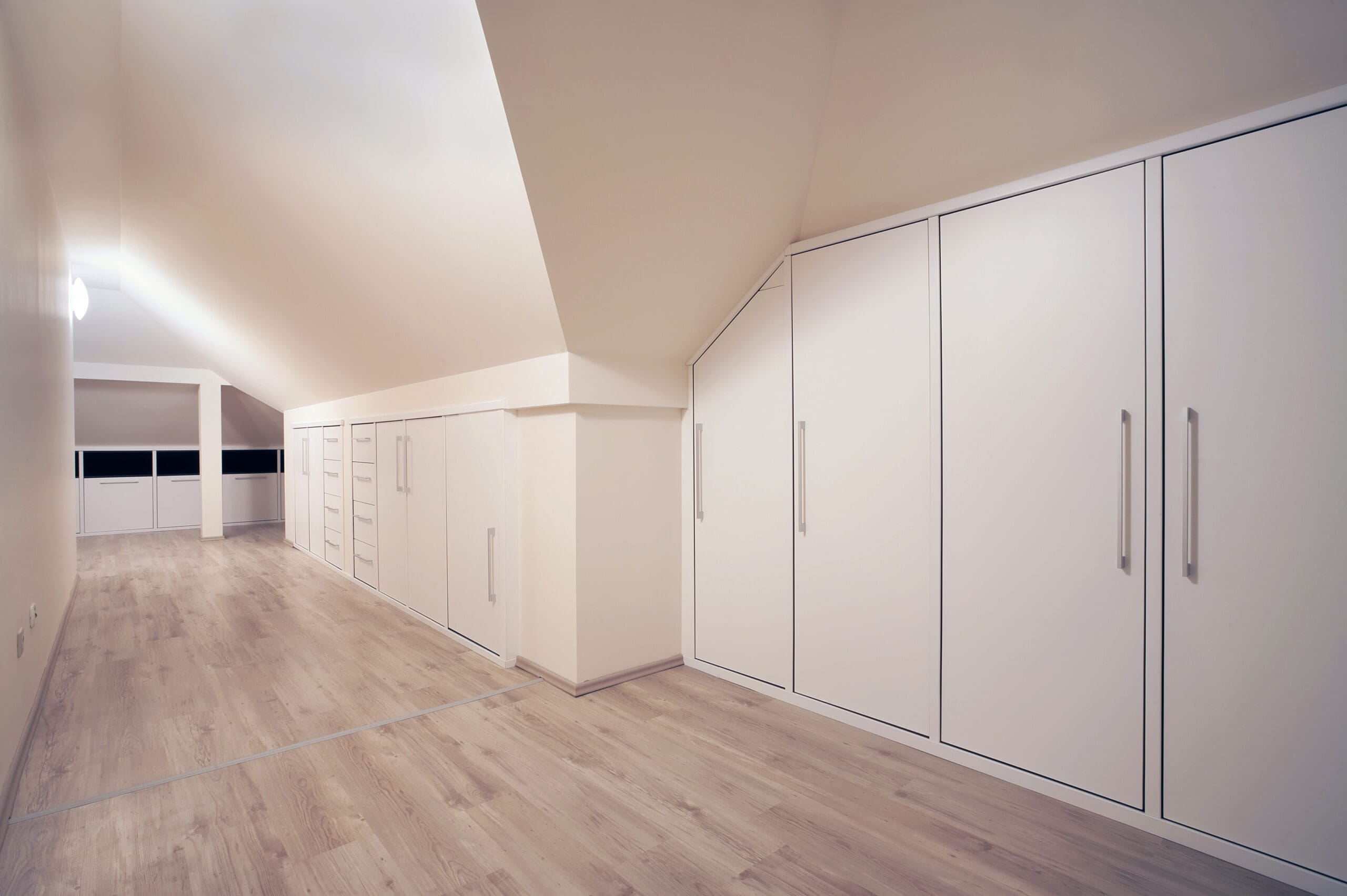 A spacious attic room with sloped ceilings and light-colored wooden flooring. Built-in cupboards and drawers line the walls, providing ample storage. The room is well-lit by ceiling fixtures and windows at the far end. The overall ambiance is clean and modern.