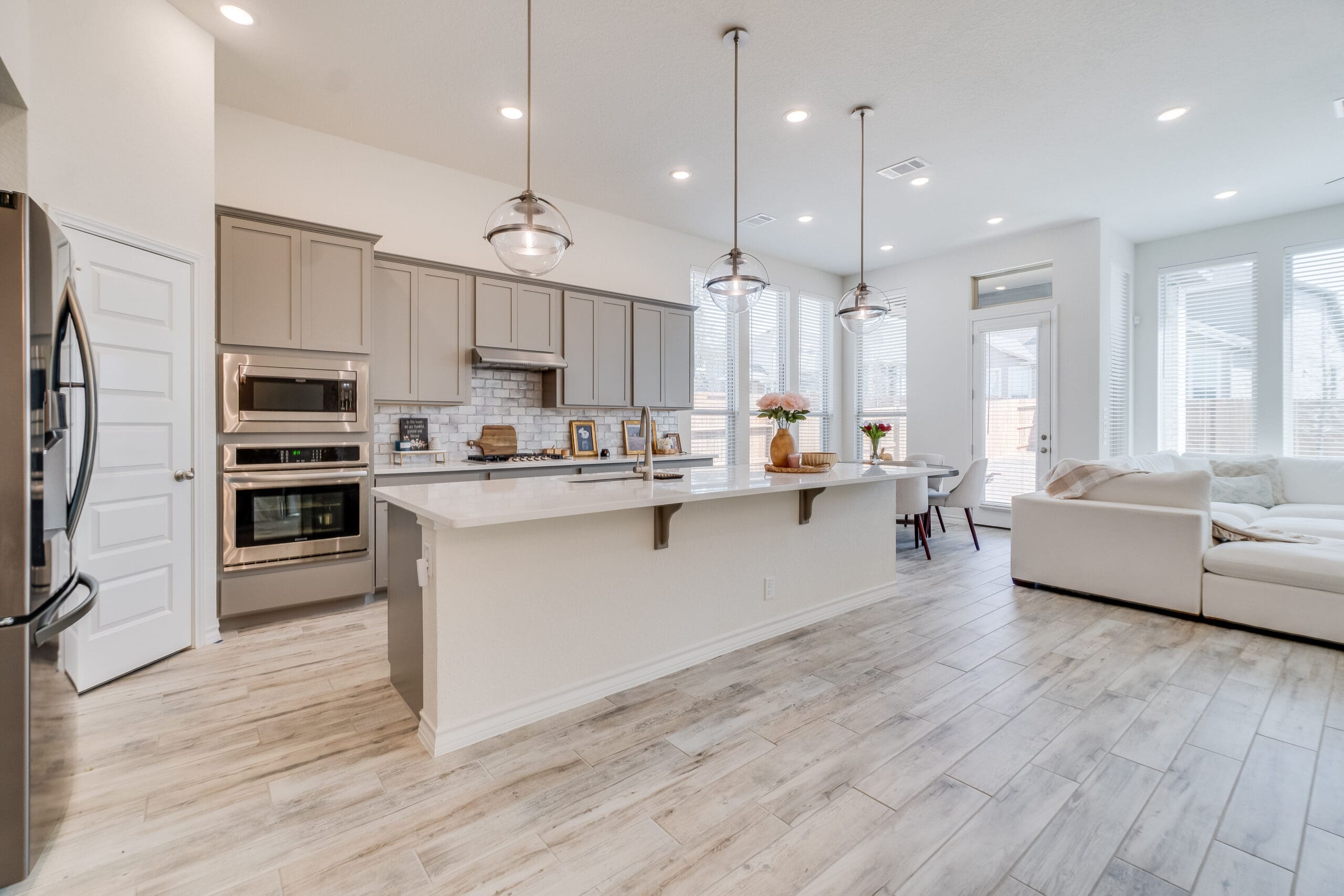 A modern open-concept kitchen and living area with light wood flooring. The kitchen features gray cabinets, stainless steel appliances, a large white island with pendant lights, and a beige sofa in the adjoining living space. Large windows provide ample natural light.