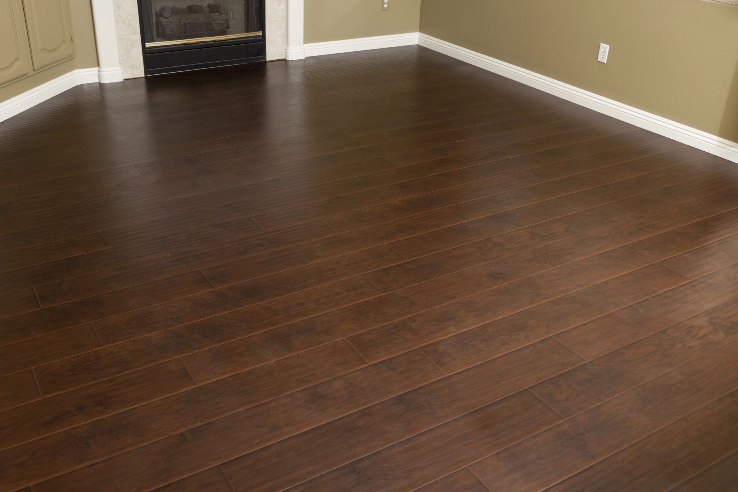 A room with dark brown wooden laminate flooring, beige walls, and white baseboards. A corner of a fireplace with a white mantle is visible in the background. The room is empty and well-lit.