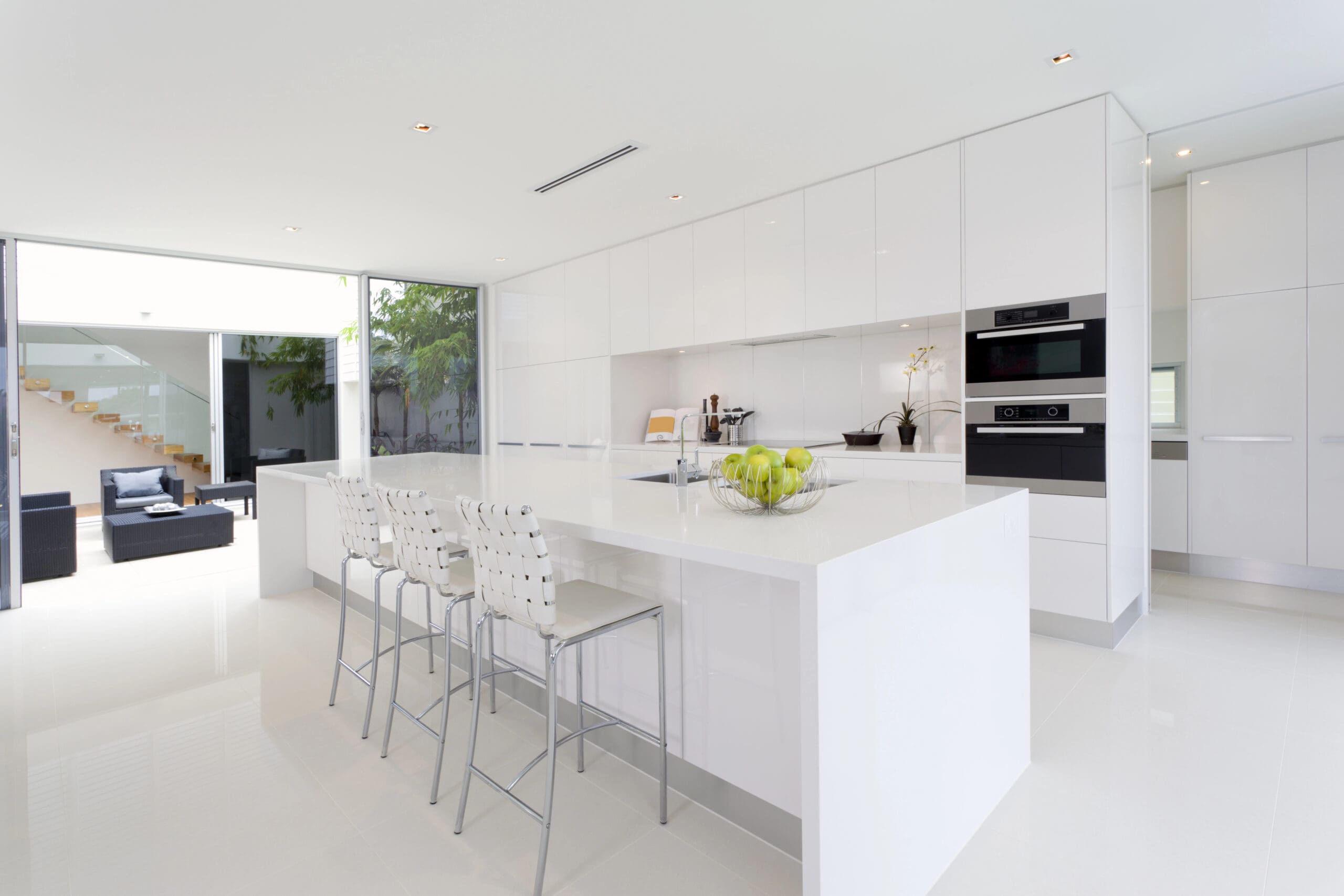 A modern, sleek white kitchen featuring a large island with four bar stools, an electric stovetop, built-in oven, and a bowl of green apples. The open design leads to a spacious living area with black couches and a staircase in the background.