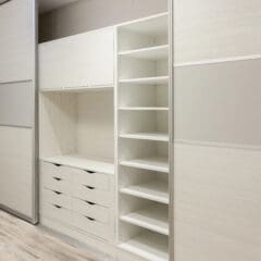 A modern closet with sliding doors, featuring a section with multiple open shelves on the right, several drawers below, and closed cabinets above. The closet has a minimalist white and light wood design, with a smooth, light-colored wooden floor.