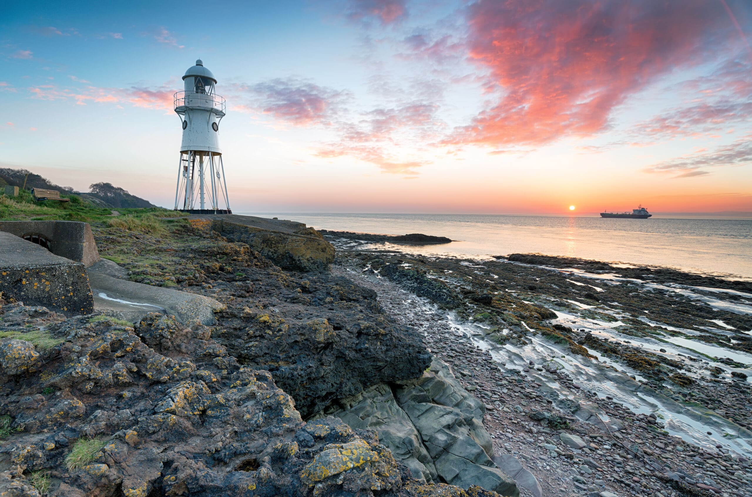 A lighthouse stands on rocky terrain by the shore under a vibrant pink and orange sunrise sky. The sea stretches towards the horizon with a cargo ship visible in the distance. The scene is serene and tranquil with rocks and shallow waters in the foreground.