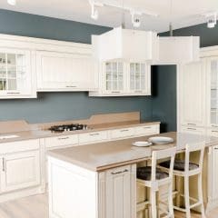 A bright, modern kitchen featuring white cabinetry and light beige countertops. An island with a beige countertop, two stools, and a sink is centered, with three square pendant lights above. The backsplash and walls are painted in a soft blue-gray hue.