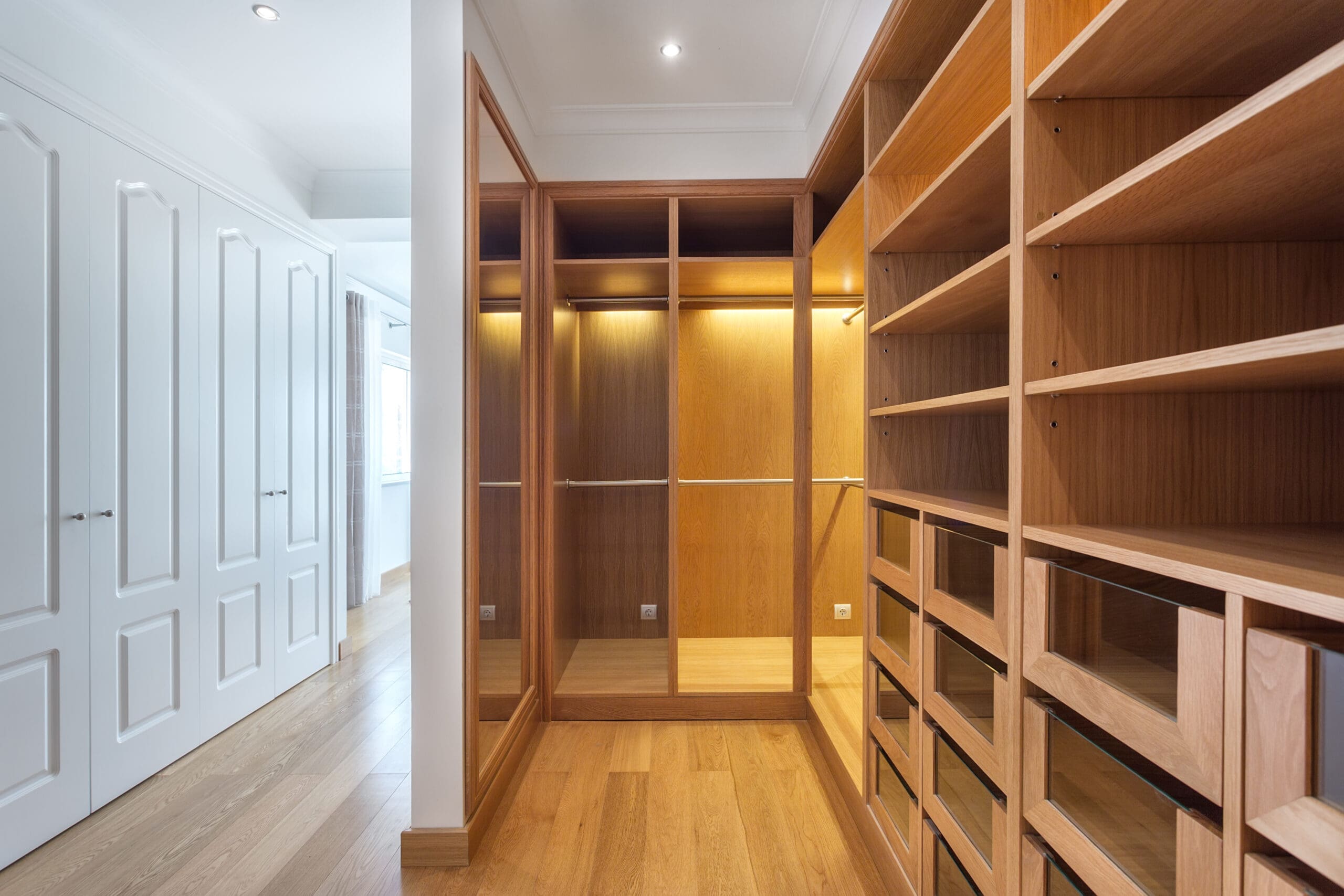 A spacious walk-in closet with wooden shelves, drawers, and hanging rods. The closet features an open design with ample storage space and natural light coming in from a window. There are white double doors on the left leading into the closet space.