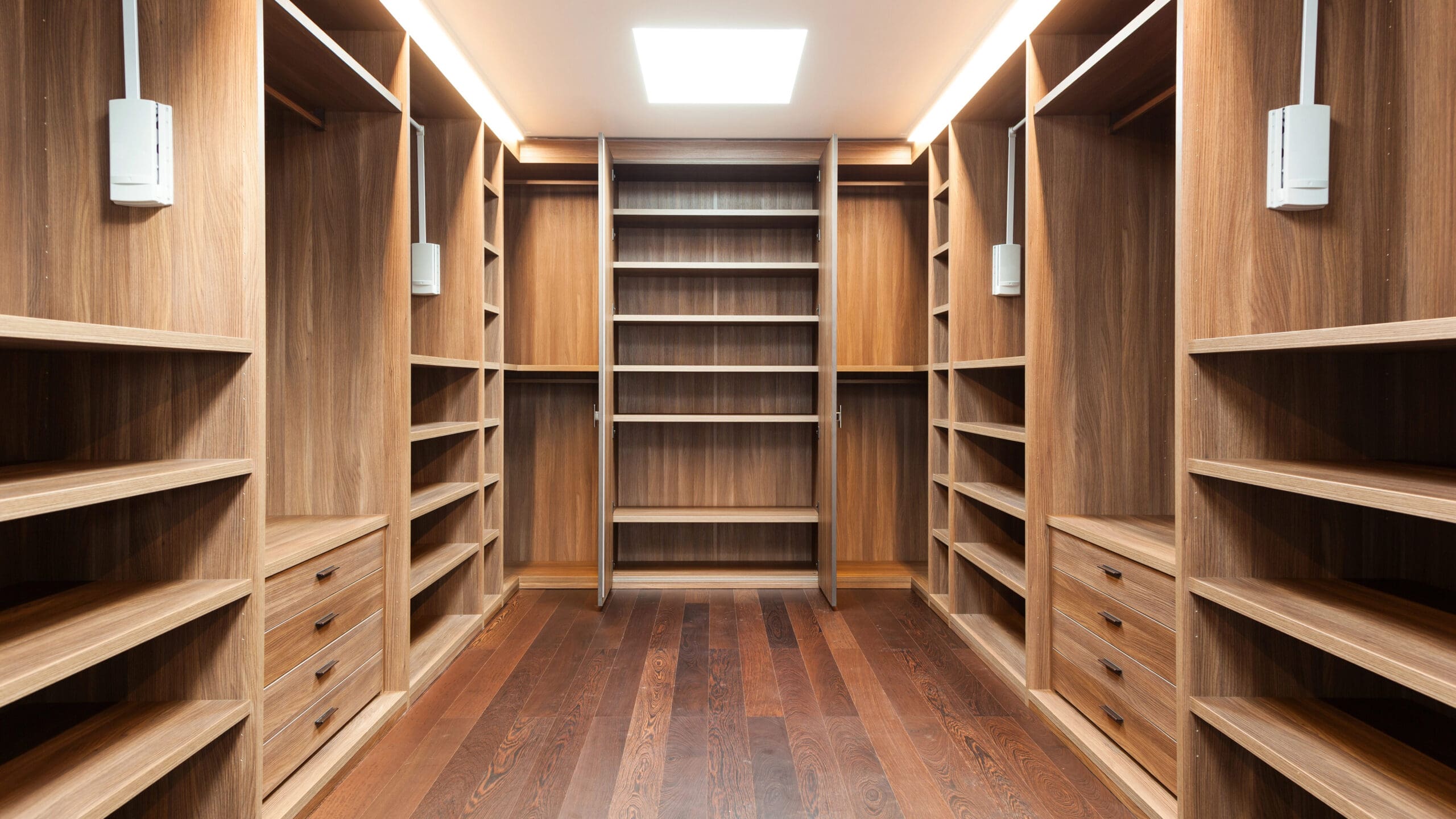 A spacious walk-in closet with wooden shelves, drawers, and hanging spaces on both sides. Bright lighting illuminates the closet, highlighting the wood grain and organized layout. A ladder-style shelving unit is centered at the end of the closet.