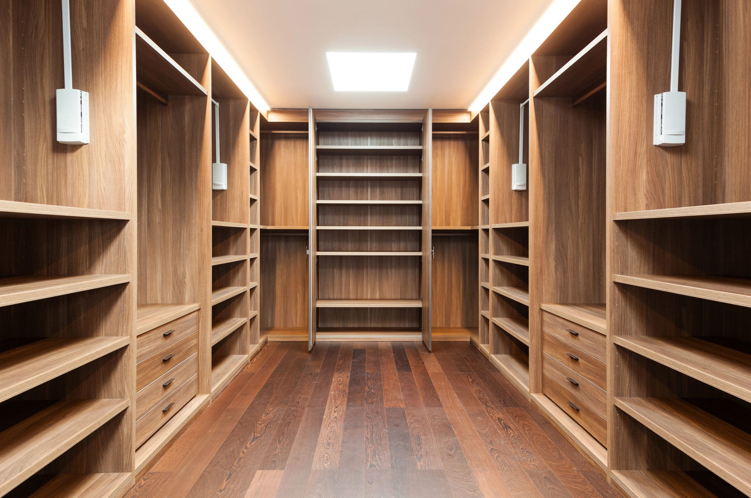 A spacious walk-in closet with wooden flooring and shelving units. The closet features an abundance of open shelves, cubbies, drawers, and hanging rods, all made of matching wood. Natural light illuminates the room from a ceiling-mounted skylight.