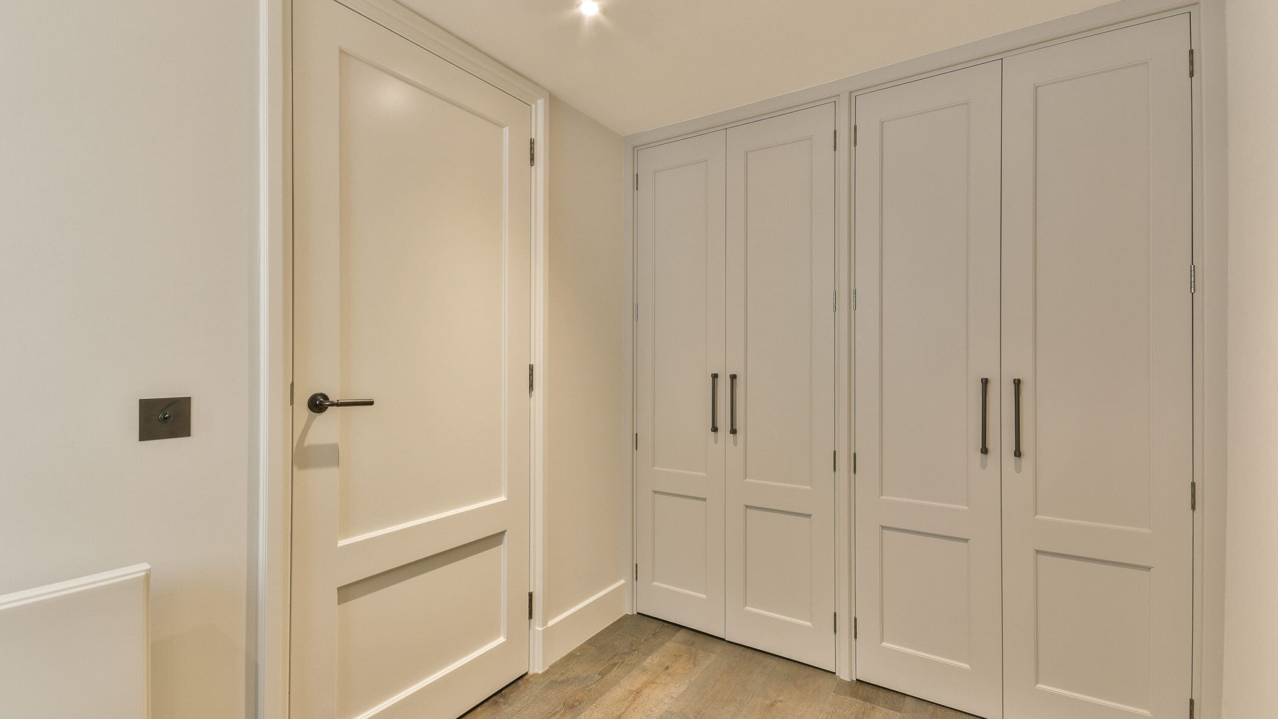 A minimalist room corner with a closed white door on the left, adjacent to double white closet doors with black handles. The walls are light-colored, and the flooring is wooden. Ceiling lights illuminate the area, indicating a clean, modern design.