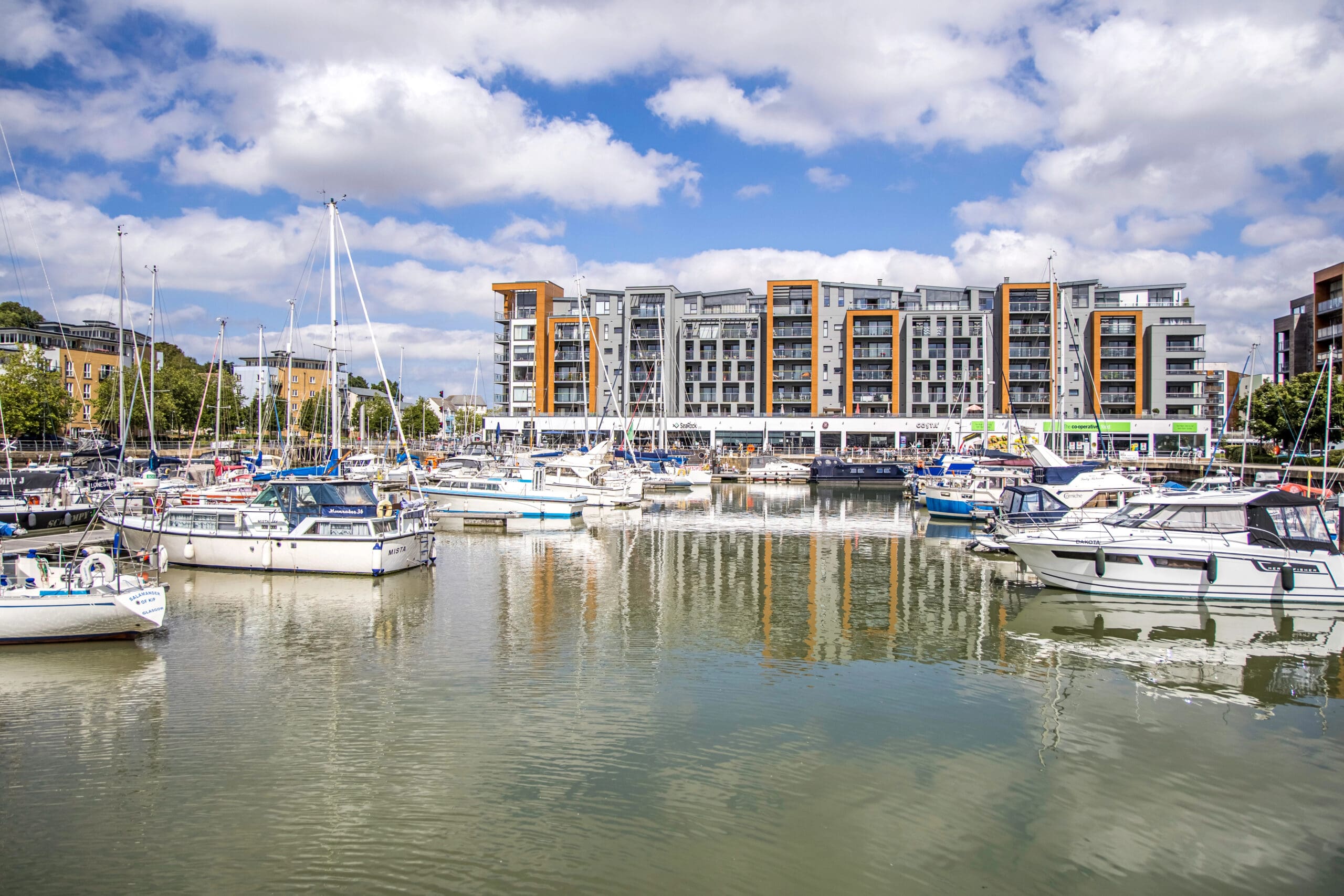 A marina filled with various sailboats and yachts docked on calm water, with modern multi-story apartment buildings in the background. The sky is mostly cloudy with patches of blue, creating a serene waterfront scene.