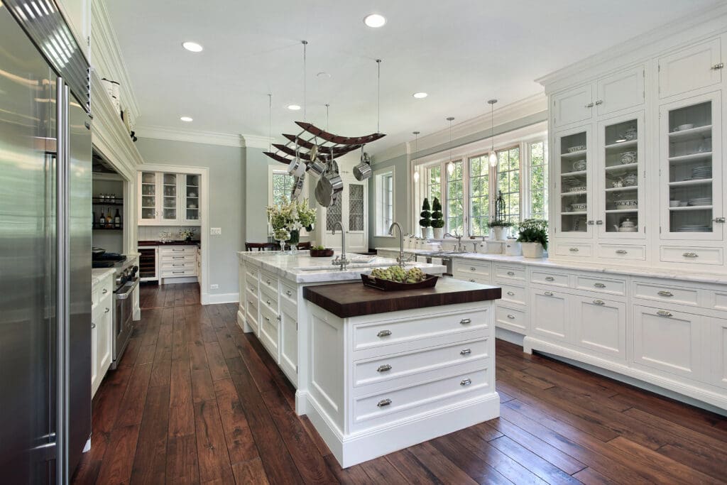 A spacious, light-filled kitchen with white cabinetry, two large island counters featuring dark wood tops, and a wooden floor. Stainless steel appliances are on the left, with a hanging pot rack above one of the islands and large windows on the right.