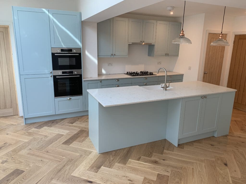 A modern kitchen with light blue cabinets, white countertops, and wooden herringbone patterned flooring. The kitchen island features a built-in sink and two hanging pendant lights overhead. Appliances include a built-in oven and a gas stovetop. A natural wood door is visible in the background.