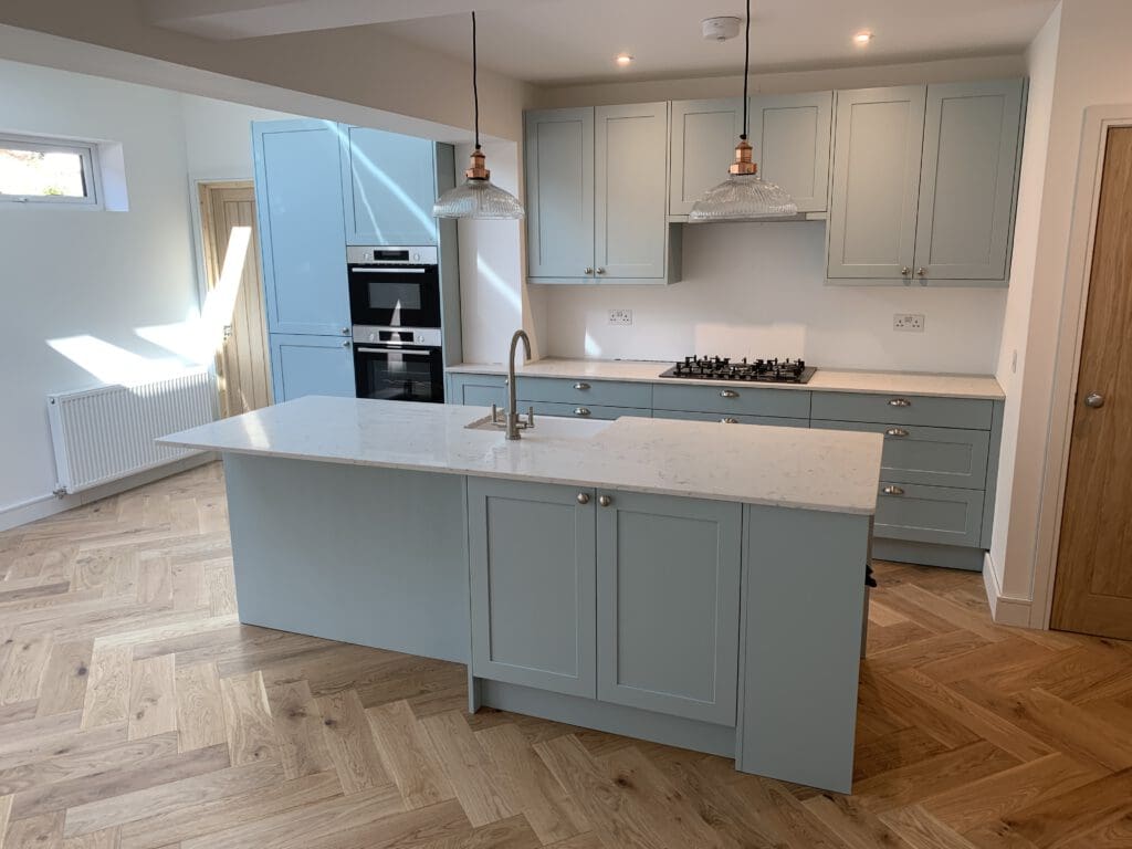 Modern kitchen with light blue cabinets and a large island with a white countertop. The island has a built-in sink and two overhead pendant lights. The kitchen features a double oven, gas stovetop, and herringbone wood flooring. A window allows natural light in.