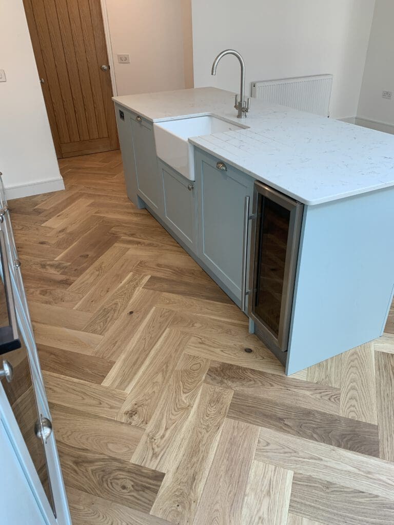 A kitchen with a light wood herringbone-patterned floor. A light blue island counter features a white farmhouse sink, pull-down faucet, and a built-in wine cooler. The white countertop contrasts with the island's color. A wooden door and radiator are visible.