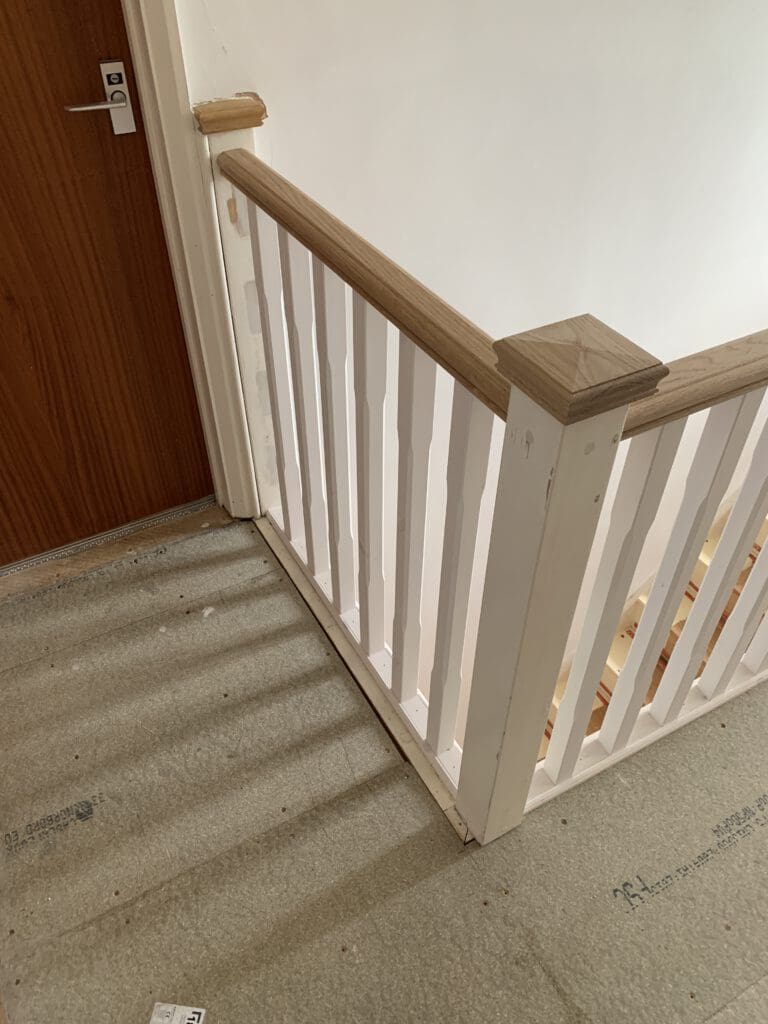 A partially painted wooden stair railing with vertical white balusters and a natural wood handrail. The railing is installed at the edge of a carpeted staircase landing. A brown wooden door is visible in the background.