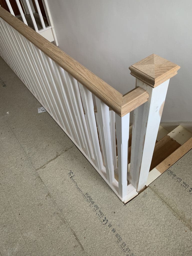 A wooden stair railing with a light wood handrail and white vertical slats is shown. The newel post at the end has a squared top. The floor appears to be under construction, with a pale, textured surface and visible measurements written on it.