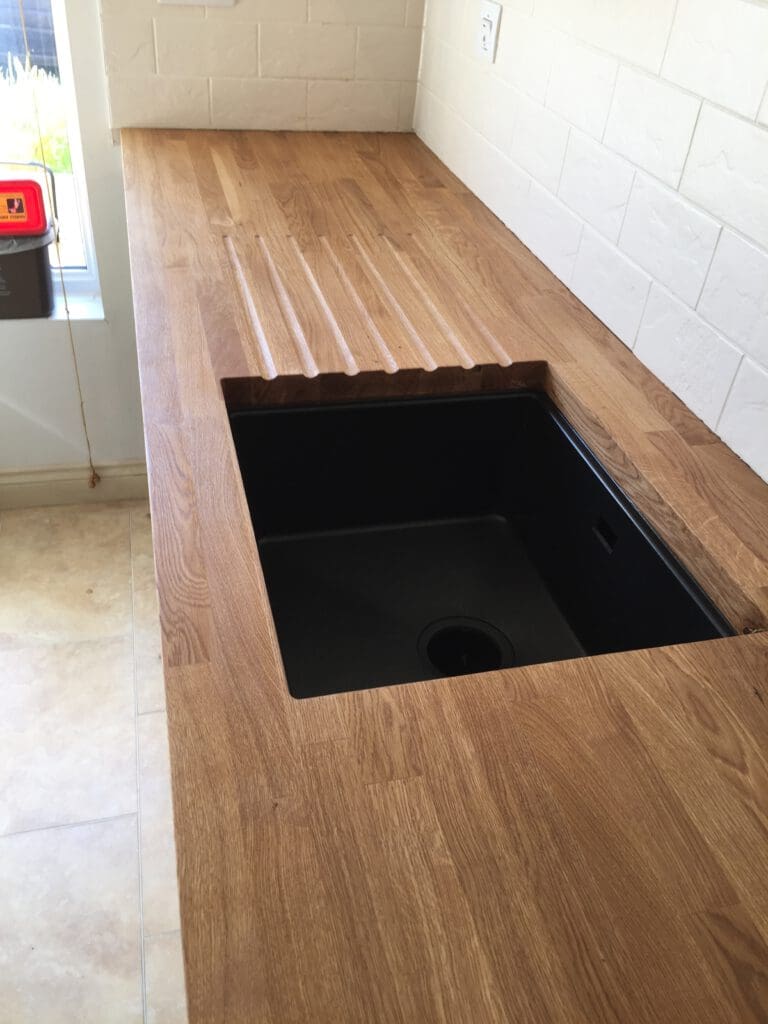 A kitchen countertop with a wooden finish features an inset black sink. Above the sink, there are grooves carved into the countertop for draining. The backdrop includes white subway tiles and a window to the left, letting in natural light.