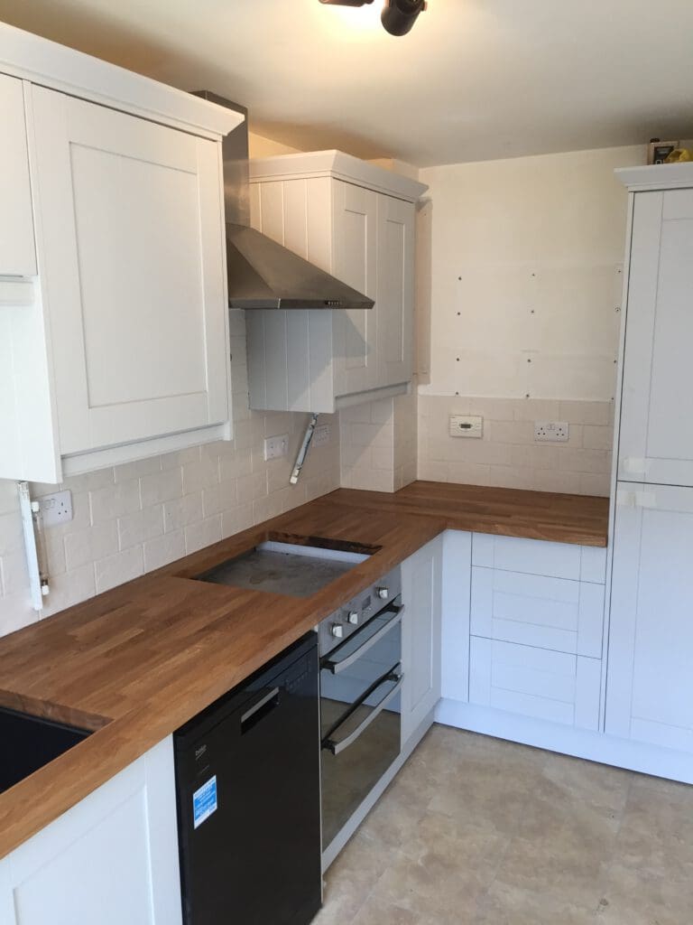 A modern kitchen featuring light-colored cabinetry, wood countertops, and a sleek stainless steel range hood. Below the counters, there is a built-in oven and a black dishwasher. The walls are tiled with white subway tiles, and the floor has large beige tiles.