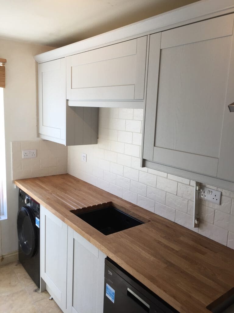 A kitchen with white cabinets and a light wood countertop. The countertop features an inset black sink and overlooks a white tiled backsplash. Below the counter, there is a washing machine and a dishwasher. The kitchen is well-lit by natural light.