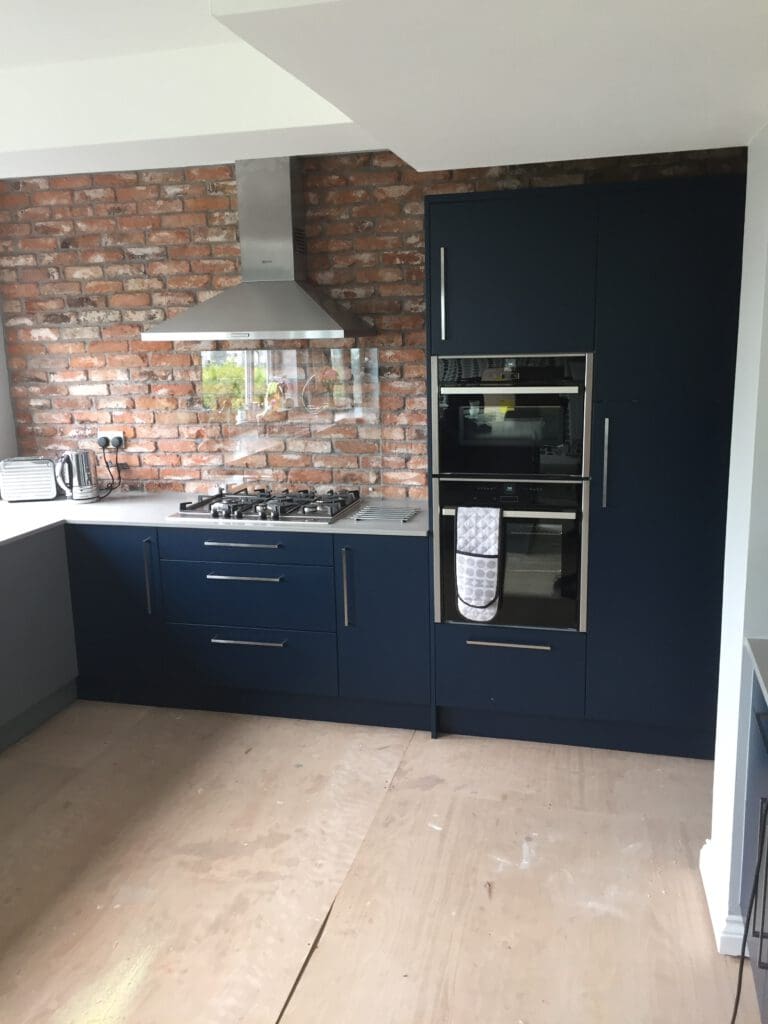 A modern kitchen featuring dark blue cabinets, stainless steel appliances, and a brick backsplash. The area includes a stovetop, oven, range hood, and built-in microwave. There's a kettle and a toaster on the countertop, and a dish towel is hanging on the oven handle.