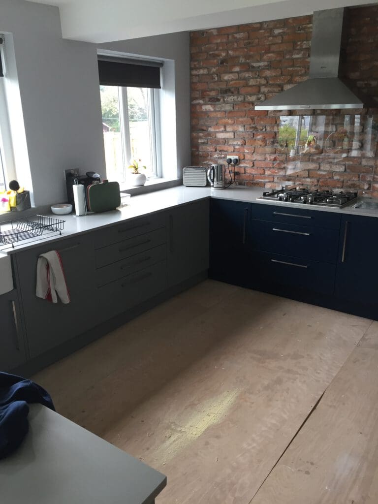 Modern kitchen featuring dark blue cabinetry, a white countertop, stainless steel appliances, and a brick accent wall with a range hood. The room is lit by a window above the sink, and a wooden floor is partially covered and unfinished.