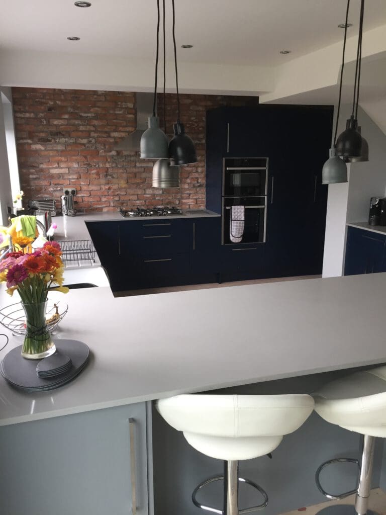 A modern kitchen with an exposed brick wall, dark blue cabinets, a built-in oven, and a gas stove. Hanging pendant lights illuminate the area. A white island countertop with white bar stools is in the foreground, with a vase of colorful flowers on the counter.