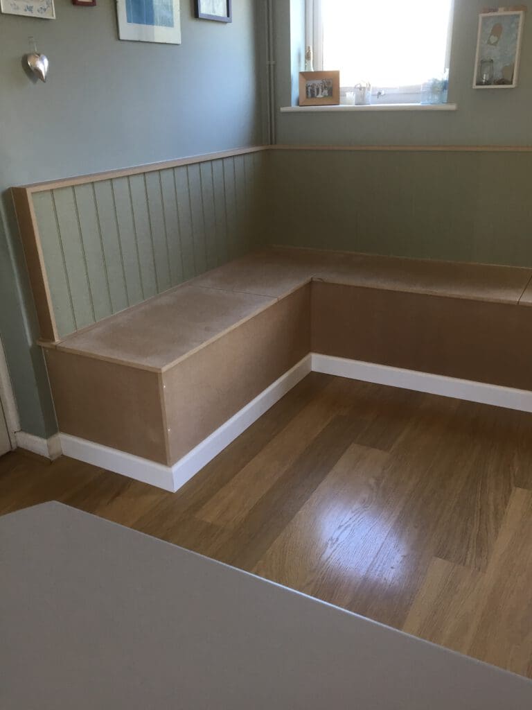 A minimalist corner seating area with light wood and green paneling. It has simple, clean lines and is positioned under a window that lets in natural light. The seating extends along two walls, creating an L shape. A few framed pictures hang on the wall above.