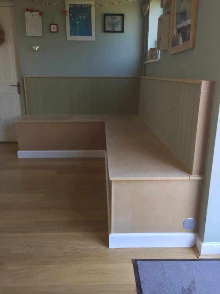 A corner of a room featuring a built-in L-shaped wooden bench with storage compartments underneath. The walls are decorated with framed pictures and a colorful garland, and the floor is light wood with a small rug in the foreground.