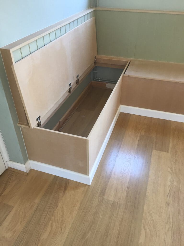 A light wood bench with a hinged seat is open to reveal a storage compartment underneath. The bench is set against a light green wall with tile accents, and the floor is made of light-colored wood.