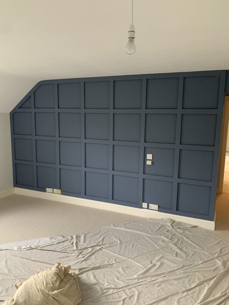 A room with a dark blue grid-patterned accent wall with moulding, a light beige carpet, and a bare lightbulb hanging from the ceiling. Against one wall, there are socket outlets and switches. A sheet covers part of the floor.