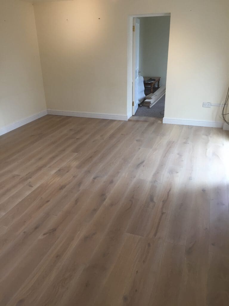 A freshly renovated room with light wood flooring and cream-colored walls. The room features an open doorway leading to another area with stacked items on the floor. The room is well-lit with no furniture visible.