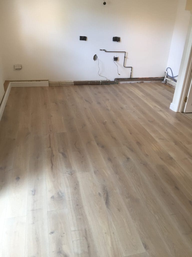 A room with light-colored wooden laminate flooring, white walls, and some exposed electrical outlets and wiring on the back wall. There are no furnishings, giving the space an unfinished and open appearance.