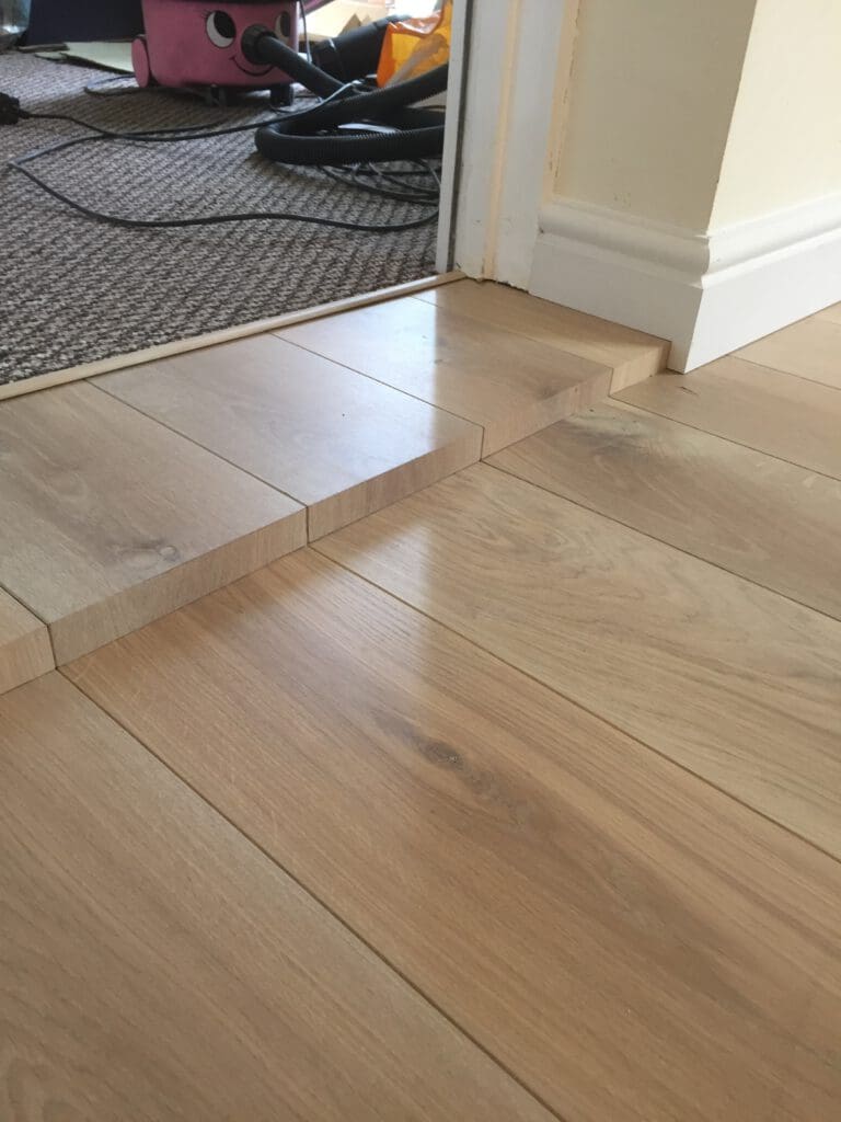 A transition in flooring is shown where a wood laminate floor meets a carpeted area. The laminate floor has been cut and staggered to accommodate the change in height between the two surfaces. A vacuum cleaner is partially visible on the carpeted side.