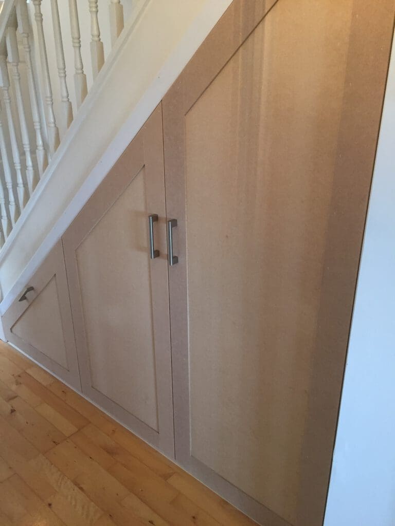 A closed storage area built into the sloped space under a staircase. The storage unit has two panel doors with silver handles and a smaller adjacent door, all made from unfinished wood. The surrounding area features a light-colored wooden floor and white walls.