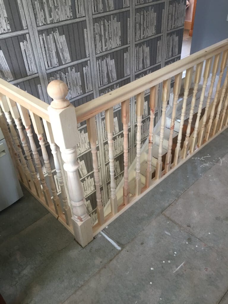 A light wood staircase banister is shown in a home under construction or renovation. The banister encloses a stairwell, and the background reveals a wall with wallpaper designed to look like shelves of books. The floor is unfinished, indicating ongoing work.