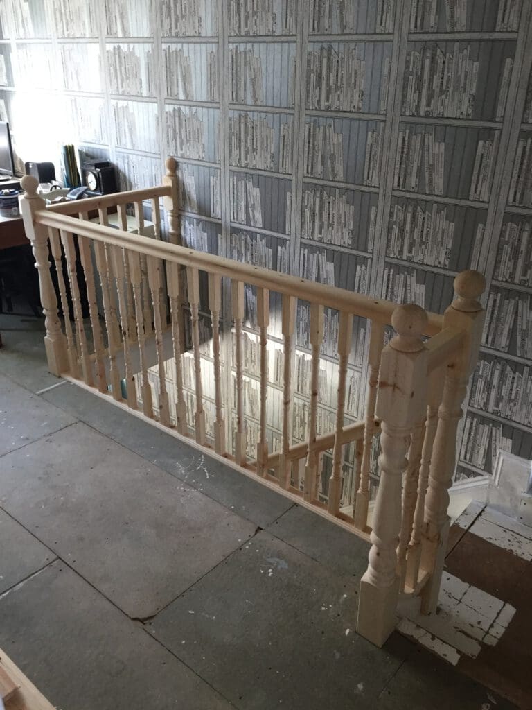 A wooden staircase railing with intricately carved balusters is partially assembled in a room. The walls are adorned with wallpaper designed to look like shelves full of books. Various construction tools and materials are scattered around the unfinished floor.