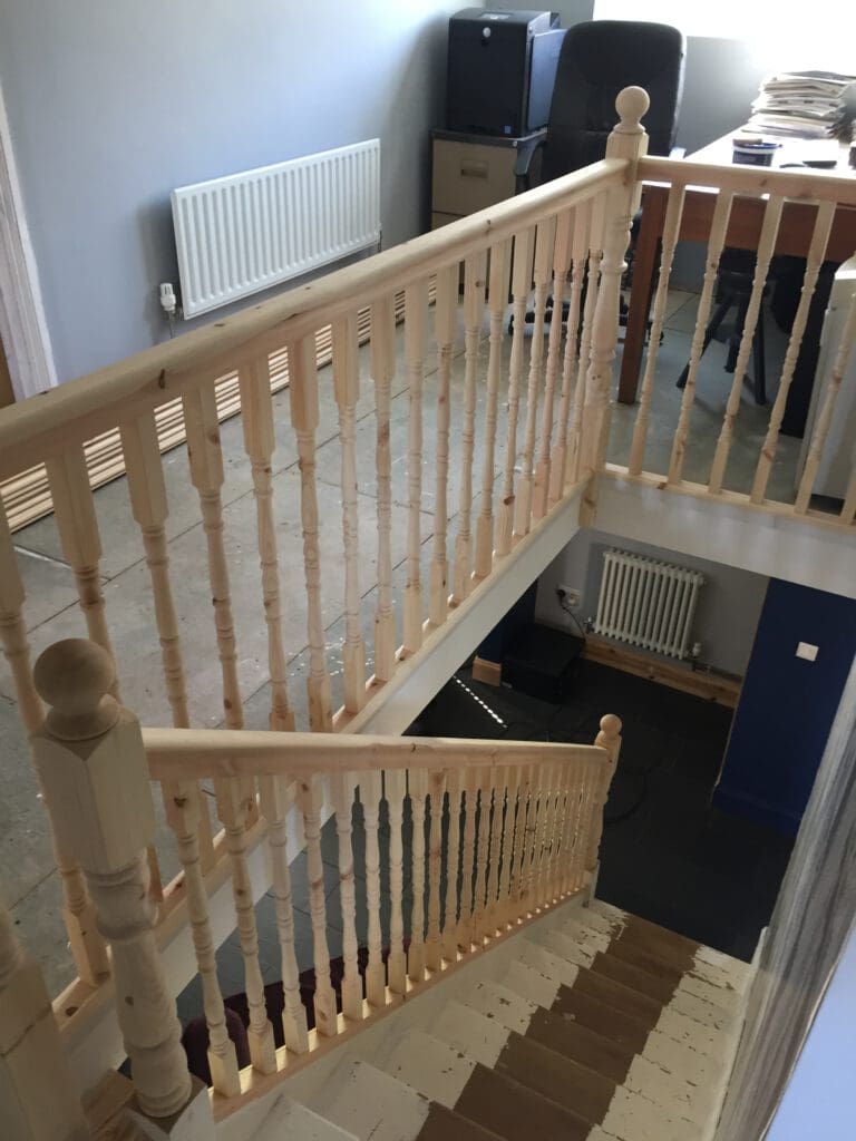 A wooden staircase with light-colored balusters and handrails leads up to a small landing area. The landing has a desk cluttered with paper and an office chair. The floor on the landing is unfinished, and a radiator is visible along the wall.