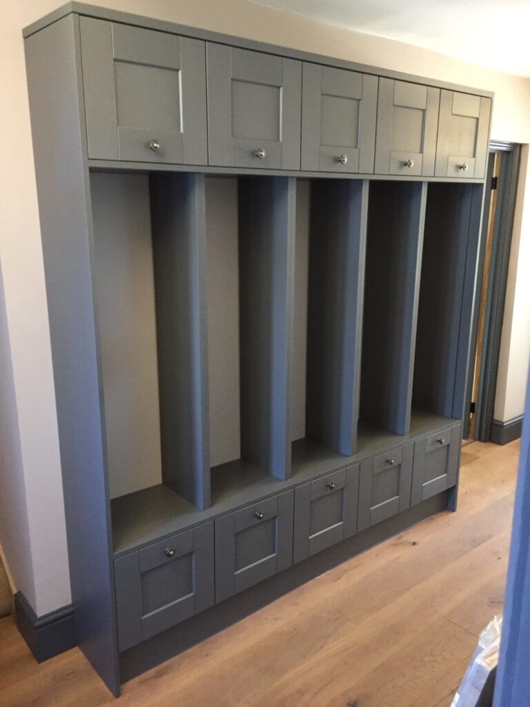 A grey built-in storage unit featuring two rows of cabinets and five vertical cubbies. The unit has upper cabinets with handles and five bottom drawers, finished with matching hardware, and is set against a light-colored wall with a wooden floor in a lit room.